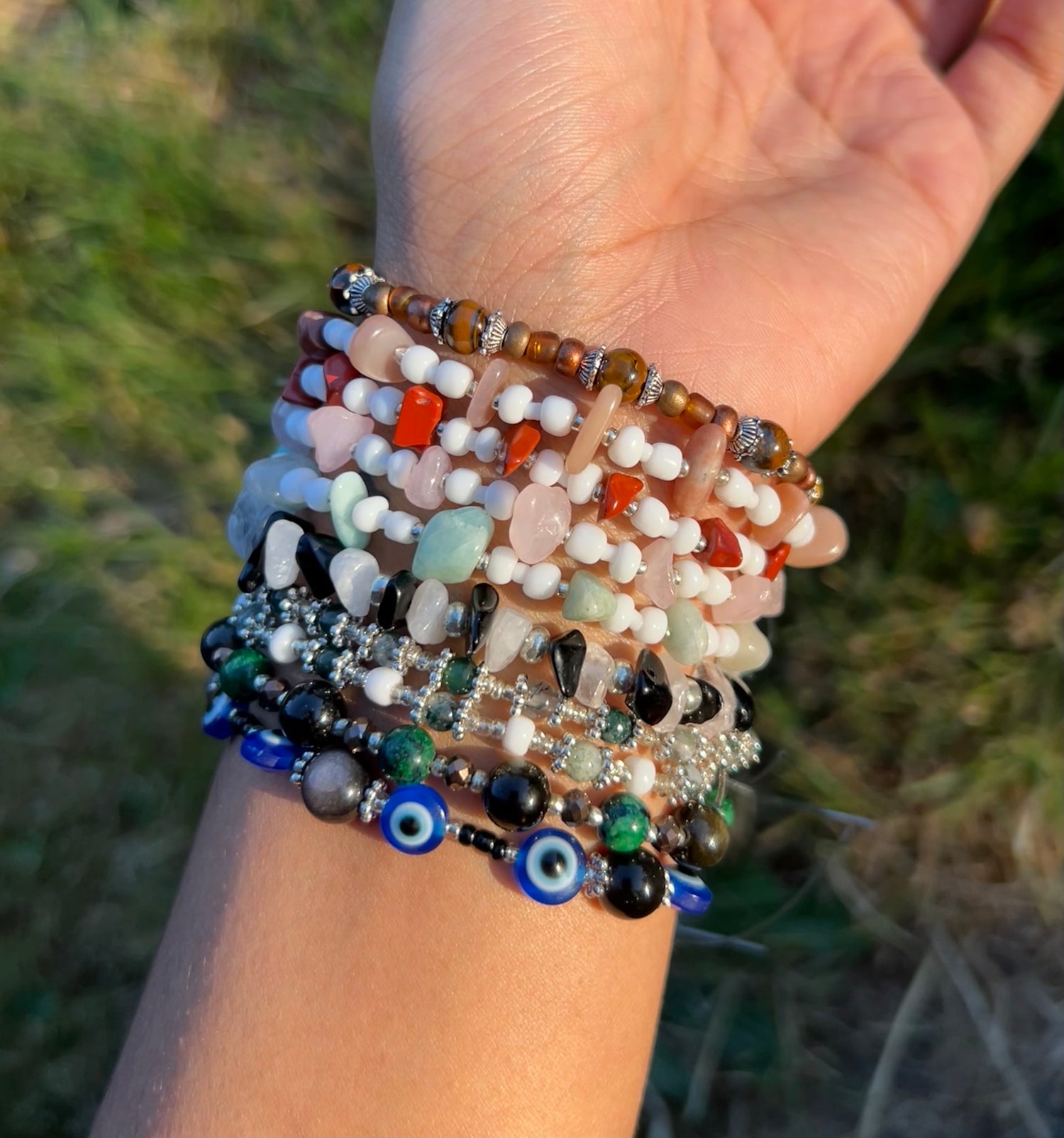 Picture of wrist with 10 different variations of crystal bracelets including evil eye bracelets and rose quartz, red jasper, obsidian, moss agate, and tigers eye beads.